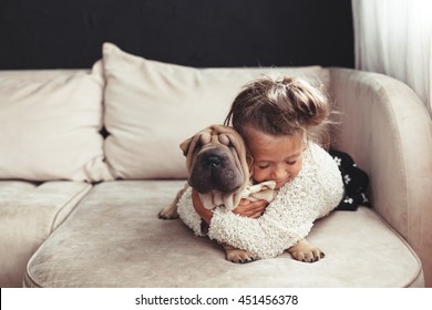 Home portrait of cute child hugging with puppy of Chinese Shar Pei dog on the sofa against black wall