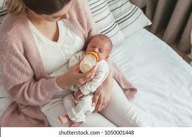 Home Portrait Of A Newborn Baby With Mother On The Bed. Young Mom Feeding Her Child From Bottle.