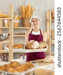 Home production of bread and pastries, work room, storage room. Young girl baker are standing near product range, hold lot of bread. Teen pupil work in baking and selling bread products