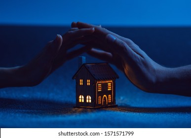 Home Protection. Male And Female Hand On A Small House In Which The Windows Glow With Warm Light