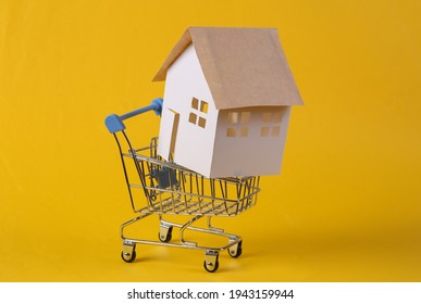 Home purchase. Mini paper house figurine in supermarket trolley on yellow background. 