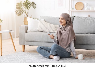 Home Relax. Smiling muslim girl in hijab unwinding with smartphone and coffee on floor in living room, enjoying weekend, free space - Powered by Shutterstock