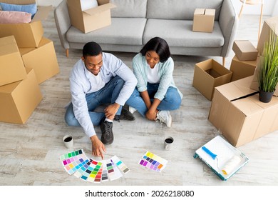 Home Renovation Concept. Young Black Couple Choosing Color Palette For Painting Walls In New House, Above View. African American Spouses Looking At Palettes While Sitting On Floor Among Carton Boxes