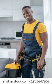 Home Repair. Dark-skinned Service Man Standing In The Kitchen And Holding A Box With Tools