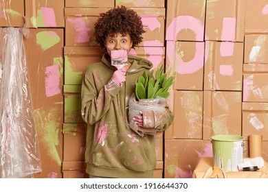 Home Repair And Makeover Concept. Curly Haired Woman In Sweatshirt Covers Mouth With Paint Brush Does Repair At Apartment Moves In New House Or Apartment Holds Potted Cactus Poses In Messy Room