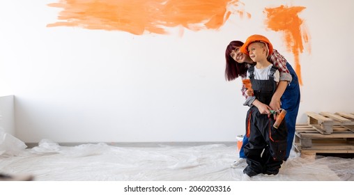 Home Repairs. Happy Family Mom And Son Painted The Wall Orange. Wall Painting. Renovation Or Remodel Room Concept. Woman And Child In Helmet On Wall Background. Banner. Place For Text.