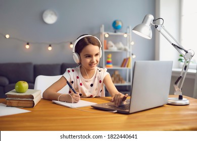 Home schooling concept. Hardworking little girl in headset watching online lesson on laptop, taking notes into her copybook indoors