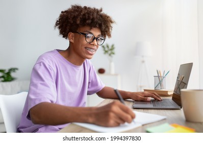 Home Schooling Concept. Smart Black Teen Guy Using Laptop At Table, Writing In Notebook During Online Lesson Indoors. Happy African American Student Participating In Web Meeting With Tutor