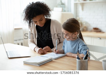 Home schooling conducted by parent or home tutor during quarantine time concept. African caring stepmom or nanny helps with school subject little daughter adorable kid girl, seated at table in kitchen