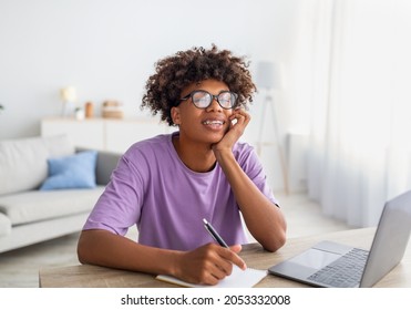 Home Schooling, Online Education Concept. Happy Black Teen Sitting At Desk With Laptop Computer, Studying Remotely, Taking Notes During Class. Cool Afro Teenager Having Lecture On Pc