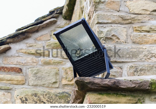 Home security light outdoor\
on the corner of a stone cottage. Metal halide floodlight\
enclosure.