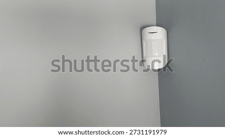 Home security system sensor mounted on a gray wall. Close-up shot of a modern indoor security device in a residential interior. Minimalist and clean design.