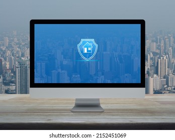 Home With Shield Flat Icon On Desktop Modern Computer Monitor Screen On Wooden Table Over Office Building Tower And Skyscraper In City, Business Home Insurance And Security Online Concept