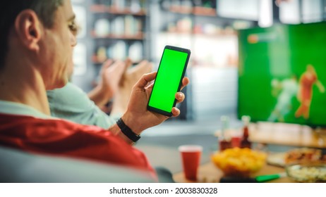 At Home Soccer Fan Holds Green Screen Chroma Key Smartphone, with Friends Watches Football Game on TV, Cheer for Favourtite Sports Team to Win Championship. Football Club Play. Over the Shoulder - Powered by Shutterstock