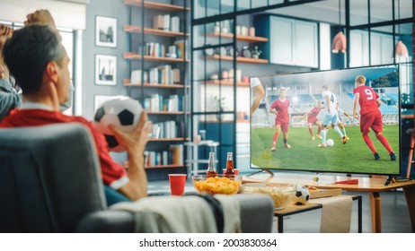 At Home Soccer Fans Sitting On A Couch Watch Football Game On TV, Cheer For Favourtite Sports Team To Win Championship. Screen Shows Professional Football Club Play. Over The Shoulder