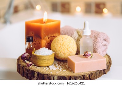 Home Spa Products On Wooden Disc Tray: Bar Of Soap, Bath Bomb, Aroma Bath Salt, Essential And Massage Oils, Candle Burning, Rolled Towel Inside Bathroom By Tub, Water Running. Cozy Relaxing Concept.