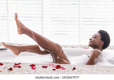 Home spa and relax on weekends. Cheerful millennial african american female in bathtub with foam and petals, raises legs and enjoys bathing in bathroom interior in morning, profile, copy space