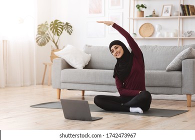 Home sport for muslim women. Smiling arabic girl in hijab practicing online yoga, watching video tutorial on laptop, excersising in living room - Powered by Shutterstock