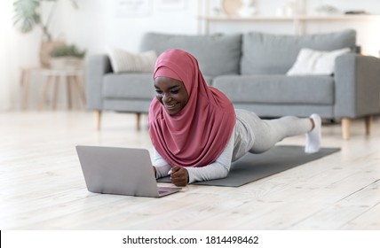 Home Sports. Active Fit Black Muslim Lady In Hijab Doing Yoga Plank Exercise In Front Of Laptop, Watching Online Tutorial Lessons On Computer, Training On Fitness Mat In Living Room, Copy Space