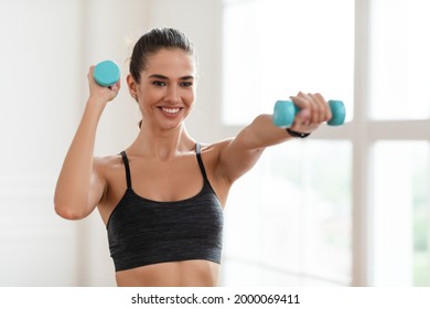 Home Sports During Covid Lockdown. Young Woman Doing Fitness Exercises With Weights Indoors, Holding And Using Dumbbells. Happy Lady Strengthening Her Arm Muscles, Leading Healthy Lifestyle