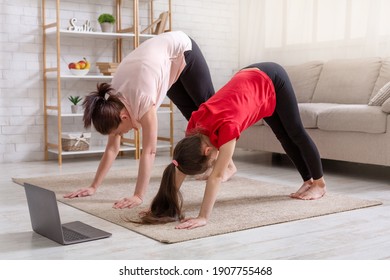 Home Sports During Covid. Young Mother And Her Daughter Exercising At Home To Online Video On Laptop, Standing In Downward Facing Dog Pose. Parent And Kid Doing Domestic Yoga