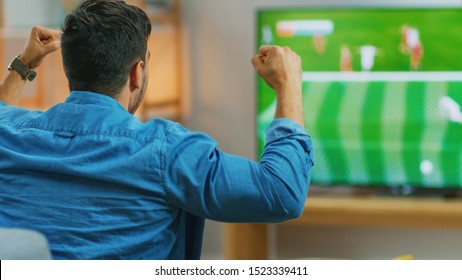 At Home Sports Fan Watches Important Soccer Match On TV, Cheering For His Team, Celebrates Doing YES Gesture After The GOAL Brings Victory To His Team. Cozy Living Room.