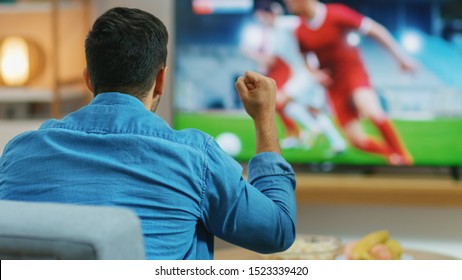 At Home Sports Fan Watches Important Soccer Match On TV, He Aggressively Clenches The Fist, Cheering For His Team. Cozy Living Room With Snacks And Drinks On The Table.