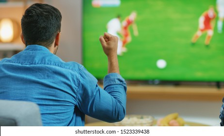 At Home Sports Fan Watches Important Soccer Match On TV, He Aggressively Clenches The Fist, Cheering For His Team. Cozy Living Room With Snacks And Drinks On The Table.