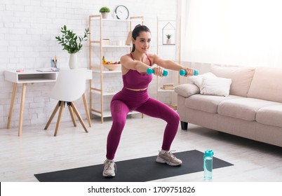 At home sports. Latin girl doing squats with dumbbells in living room