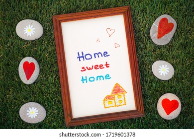 Home Sweet Home Picture In Wooden Frame  On Green Grass Background With Some Pebbles With Daisies And Hearts.