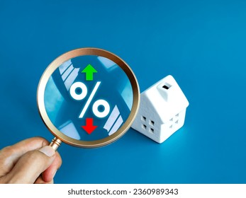 Home tax concept. Real estate, land and property annual taxation. Percentage icon with green up and red down arrow and white house model in magnifying glass lens, hold by hand, on blue background. - Powered by Shutterstock