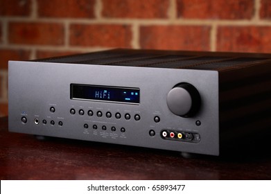 Home Theatre Amplifier A High End Hi-Fi Home Theatre Amplifier With A Red Brick Wall Backdrop. Black Enclosure With Blue LCD Display.