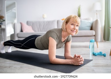 Home Training Concept. Smiling Senior Woman Doing Elbow Plank On Yoga Mat In Living Room. Positive Mature Lady Doing Her Workout Routine, Exercising Indoors, Strengthening Her Core Muscles