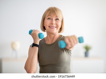 Home training concept. Strong senior woman doing exercises with dumbbells indoors. Cheerful mature lady working out her arm muscles, keeping fit, leading healthy lifestyle during covid-19 isolation