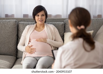 Home visit of obstetrician. Young female on last months of pregnancy sit on sofa talk to attending gynecologist checking her health state. Future mom prepare for childbirth discuss details with doctor
