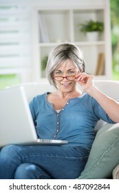 At home. A woman in her forties surfing on the Internet with her laptop on knees, she is sitting in a armchair. she laughs. she has glasses.  - Shutterstock ID 487092844