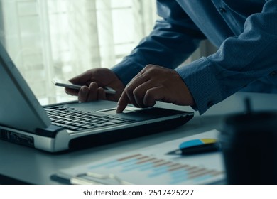 Home working concept. Close-up of businessman working on laptop while holding smartphone. With documents and charts on a table in a modern office, coding, meetings and financial spreadsheets. - Powered by Shutterstock