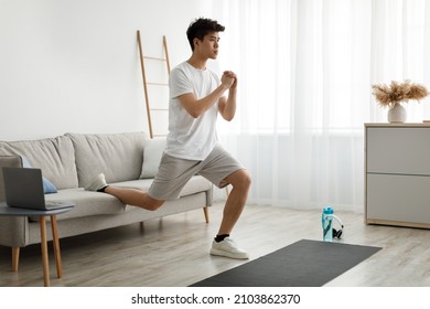 Home Workout Concept. Focused Determined Young Asian Man Doing Single Leg Split Lunge Near Sofa Exercising Using Laptop Computer During Workout At Home. Domestic Male Fitness And Sporty Lifestyle