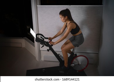 Home Workout Indoor Stationary Bike Asian Girl Biking Screen With Online Classes Woman Training On Smart Fitness Equipment Indoors For Cycling Exercise. Late At Night In Bedroom.