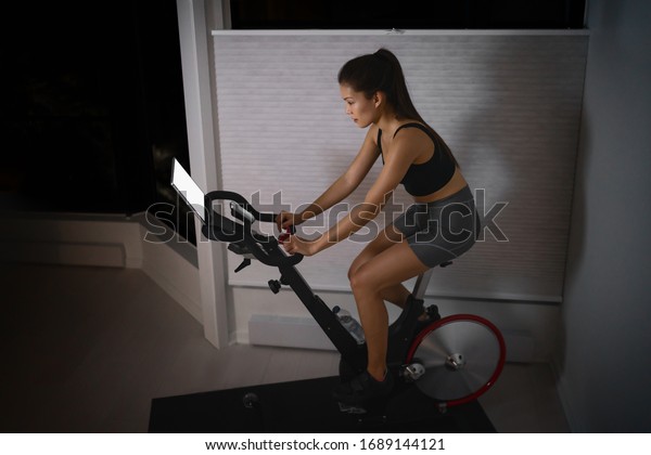 Home workout\
indoor stationary bike Asian girl biking screen with online classes\
woman training on smart fitness equipment indoors for cycling\
exercise. Late at night in\
bedroom.