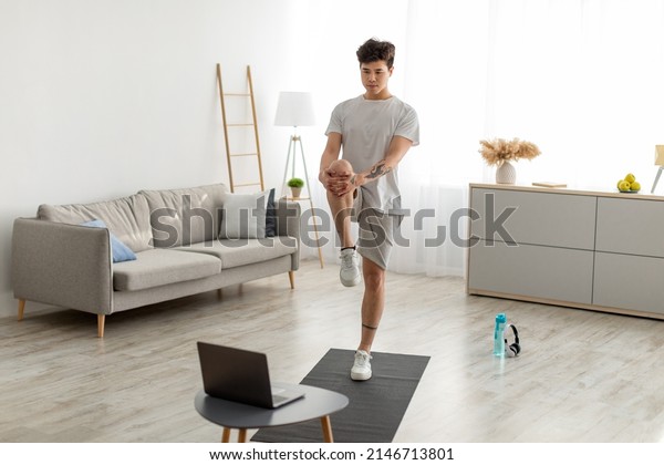 Home Workout Routine. Focused Fit Asian Guy\
Lifting Leg Up To Chest Stretching Hamstring Muscles, Doing\
Standing Knee-To-Chest Exercise, Using Pc In Living Room Indoors.\
Healthy Lifestyle Concept