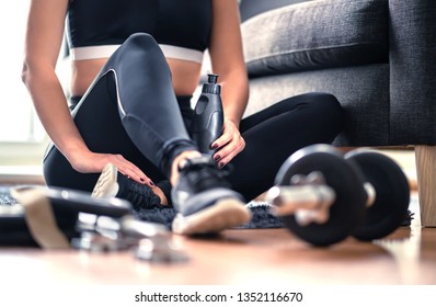 Home Workout, Weight Training And Fitness Exercise Concept. Woman In Sportswear Sitting In Living Room With Gym Equipment And Dumbbell Holding Water Bottle. Healthy Sporty Lifestyle. Dedicated Trainer