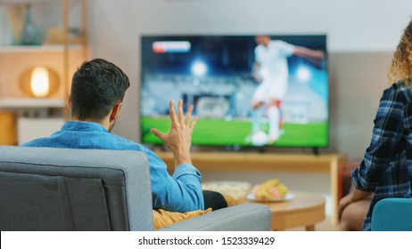 At Home Young Couple Watches Tense Moment Of Soccer Match On TV, They Worry, Guy Trying To Be Patient And Aggressively Showing Palm. Back View Shot.