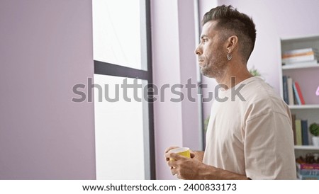 At home, a young hispanic man, expression full of sadness, standing forcelessly by the window, clutches onto his morning coffee cup, his woes clear as he looks through.