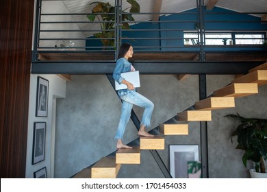 At home. Young woman going upstairs, holding laptop under arm - Shutterstock ID 1701454033