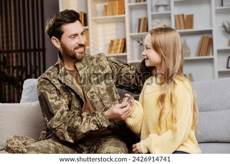 Homecoming soldier in Ukrainian uniform finds solace on living room couch, gifting his little girl his military tag, gesture of their unbreakable bond. Father and daughter look at each other with love