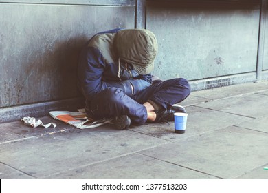 Homeless Adult Man Sitting On The Street In The Shadow Of The Building And Begging For Help And Money. Problems Of Big Modern Cities. Indifference Of People. Social Issues.