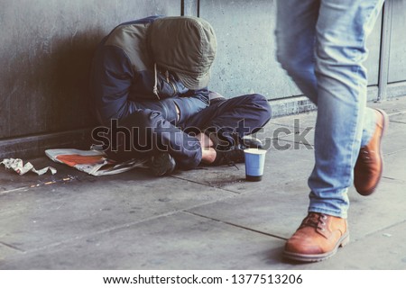 Homeless adult man sitting on the street in the shadow of the building and begging for help and money. Problems of big modern cities. Indifference of people. Social issues.