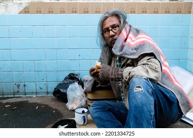 A Homeless Asian Old Man With A Long Beard Sits On The Street Eating Bread After Receiving A Delicious Snack. From The People Passing By The Poor Have No Homes On The Streets.