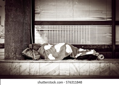 Homeless Barefooted Woman Sleep On The Urban Street In The City Sidewalk Near Building